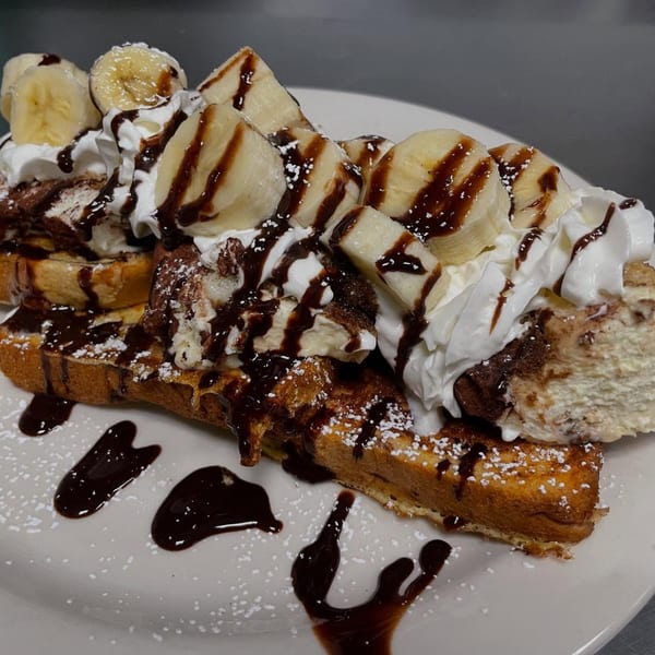 French toast topped with whipped cream, banana slices, chocolate syrup, and powdered sugar.
