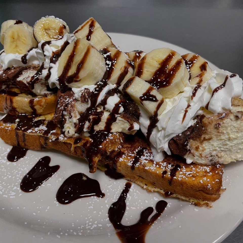 French toast topped with whipped cream, banana slices, chocolate syrup, and powdered sugar.